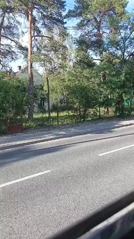 This media file captures a view from a moving vehicle, likely a car or bus, on a street in Konstancin-Jeziorna, Poland. The primary subjects are a residential building, trees, and the road. The building appears to be a multi-story house with a light-colored facade, visible through a dense arrangement of trees. Some windows and what might be a satellite dish are discernible on the house.

The scene is dominated by lush greenery, with tall pine trees and other deciduous trees lining the property. A chain-link fence and a section of a gate are visible along the edge of the property, separating it from the sidewalk and road. The sidewalk is paved with light-colored tiles, and the road in the foreground is asphalt with white lane markings. Shadows cast by the trees and the vehicle suggest it is daytime, likely late morning or early afternoon, with sunny weather. The overall atmosphere is serene and natural, typical of a suburban or semi-rural setting. No people are visible in the frame. There is no visible text in the media file.
