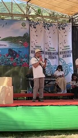 A man wearing a cap, glasses, a light-colored t-shirt, and khaki pants stands on a stage, holding a microphone in his right hand and a phone in his left. He appears to be speaking into the microphone. He is positioned in front of a banner that reads "TAMAN NASIONAL KARIMUN JAWA" multiple times. To his left are two stacked beige boxes. Behind him, to the right, two women are seated, one of whom is looking at her phone. Another man stands to the far right, also looking at something in his hands. The stage is covered with a red carpet and has a green cloth draped along the front. In the background, there is a larger banner with an image of a body of water, a diver, and plants. The stage is outdoors under a roof structure, with trees and a hillside visible in the distance. The ground in front of the stage is covered in grass, with a blue water bottle and some items on a table in the foreground.