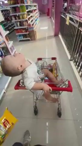 A man pushes a miniature shopping cart through a grocery store aisle. A young child, around one or two years old, sits in the shopping cart, facing away from the person pushing. The child has short, light hair and is wearing a white t-shirt and patterned shorts, and is barefoot. The aisle is brightly lit with white tile floors and pink columns and trim. The shelves are stocked with various products. The man is wearing blue jeans and gray sneakers.