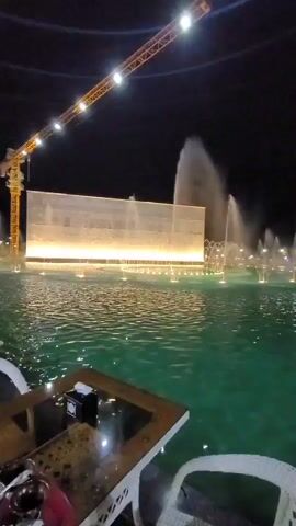 This media file depicts an outdoor setting at night. The foreground features a table with white wicker chairs, suggesting a seating area for patrons, possibly at a restaurant or cafe. On the table, a tissue dispenser and what appears to be a shisha pipe are visible.

The midground and background are dominated by a large body of water with numerous fountains actively spraying water into the air. The water is illuminated, giving it a vibrant green hue. Beyond the water, a large, illuminated rectangular structure stands, possibly a building or a decorative wall, with bright lights across its lower section. A tall, yellow construction crane with lights is positioned to the left, extending into the dark sky. The overall scene is lively and visually dynamic due to the illuminated fountains.

The activity taking place is the operation of a large fountain display, likely intended as entertainment or ambiance for visitors. The time of day is clearly night, indicated by the dark sky and the artificial lighting of the scene. There are no people clearly visible in the frame. The presence of the construction crane hints at ongoing development or construction in the vicinity. The visual elements create an atmosphere of entertainment and possibly relaxation, especially with the nighttime illumination and water features. No discernible text is visible in the media.