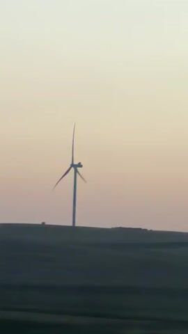 A video captured from a moving vehicle shows a solitary wind turbine in a field under a pale, dusky sky. The turbine is silhouetted against the horizon, with its blades appearing to turn slowly. The foreground consists of dark, blurred fields, indicating the speed of the vehicle. In the later frames, trees partially obscure the view of the turbine. The overall impression is one of a vast, open landscape at dusk.