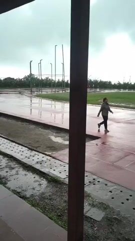 A person is running on a red track outdoors on a cloudy day. The track is wet, indicating it may have rained recently or is raining. In the background, there is a green field, trees, buildings, and a tall tower. There are also spectators seated in a covered area to the right.