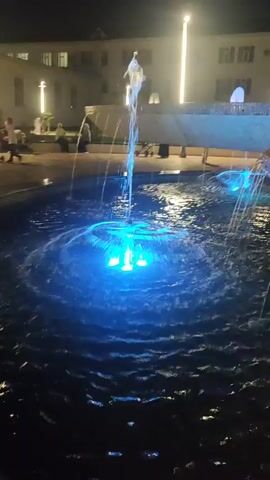 This is a short video clip taken at night, showcasing a beautifully lit fountain. The fountain is the central subject, with multiple jets of water shooting upwards. The water is illuminated by vibrant blue and purple LED lights, creating a mesmerizing visual effect as the water cascades and creates ripples on the surface of the pool. In the background, a building with several illuminated windows can be seen, suggesting an urban or semi-urban setting. The time of day is clearly night due to the darkness and the artificial lighting of the fountain and building. The weather appears calm, as the water in the fountain is not being disturbed by strong winds. There are no people clearly visible in the foreground, though there may be figures in the background, indistinct due to the low light and distance. The scene evokes a sense of tranquility and modern aesthetic, highlighting the beauty of water and light in a nighttime environment. No text is visible in the video.
