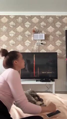 The video shows a woman and a cat in a bedroom in Belozernyi, Russia. The woman is lying on a bed, facing a large television screen. She is wearing a pink long-sleeved shirt and has her hair tied up in a bun. A grey cat is lying next to her on the bed.

The television screen displays a sequence of images, appearing to be from a movie or TV show. Some of the on-screen text is visible, including "EMMA D'ARCY" and "YOKK." The room is decorated with patterned wallpaper. A Wi-Fi router and electrical outlets are mounted on the wall above the television. The overall impression is of a relaxed home environment, with the woman and her pet enjoying entertainment. There are no specific weather or time of day cues, but the indoor lighting suggests it could be daytime or evening. The woman's posture and the presence of the cat suggest a comfortable and possibly cozy atmosphere.