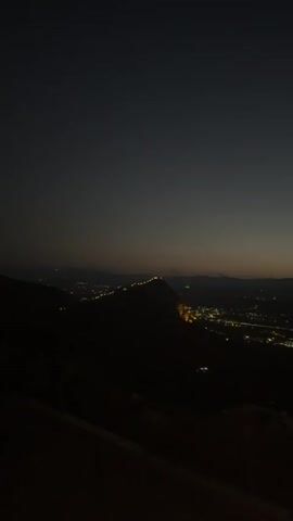 A wide shot taken at dusk or night shows a sprawling city spread out in a valley below a dark mountain range. The sky is a deep gradient of dark gray to a lighter gray near the horizon. The city lights twinkle like scattered diamonds, illuminating buildings and streets. A prominent hill or mountain ridge in the middle ground is lit by a line of lights, possibly streetlights or lights from buildings along its crest. The foreground is dark and indistinct, with what appears to be a slope or ledge. The overall impression is one of a vast, illuminated urban landscape against a dark, natural backdrop.