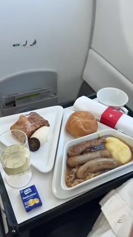 The video shows an in-flight meal service on an airplane. The meal tray contains a main course of sausages with mashed potatoes and onions, a bread roll, butter, and a dessert of strudel with ice cream. There is also a glass of white wine. In the background, the airplane cabin is visible, including seats and a window. The video then transitions to a view out the airplane window, showing a clear blue sky with clouds and land below.