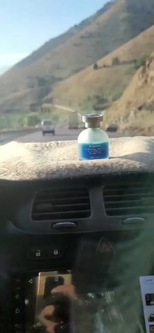 The video shows a car's dashboard with a small bottle of blue liquid with a label that says "Aroma" placed on top. The bottle is in the foreground. Outside the car's windshield, a highway can be seen with mountains in the background. Several vehicles, including trucks and cars, are driving on the highway. The sun is shining, and the sky is blue. The video appears to be filmed from the perspective of a passenger in the car.