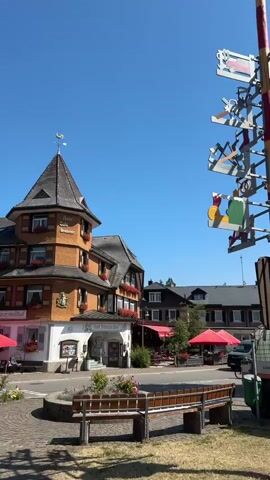 This media file captures a sunny day in Hinterzarten, Germany, likely during daytime given the bright sunlight and clear blue sky. The scene is a charming village street, possibly a town square, with traditional German architecture.

Prominently featured is a colorful, ornate signpost adorned with various cutout figures, suggesting local crafts or cultural motifs. Beside it, a wooden bench offers a place to rest. In the background, a multi-story building with a distinctive turret and red flower boxes stands out, identified by signage as "Hotel Café Schwarzwaldhof" and "Adlerweg." Other buildings with characteristic steeply pitched roofs are visible, some with red umbrellas shading outdoor seating areas, hinting at cafes or restaurants.

Vehicles are present, including a white van with a graphic logo, a smaller silver van, a utility vehicle with a water tank, and cars parked along the street and in a lot. A bicycle is also visible. A man in a patterned shirt and shorts is walking away from the camera.

Traffic signs are clearly visible, including a parking sign indicating "90 Min." and specific hours of operation "8-18h," along with directional arrows. The overall atmosphere appears peaceful and inviting, characteristic of a European village on a pleasant day.