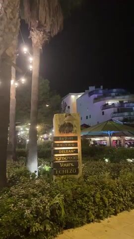 A signpost with illuminated text is visible at night. The sign has the words "MARINA VILLAGE" at the top, followed by "Pavion", "Retro", "Delean*", "Highscream", "Yours.", and "Echezo". The signpost is surrounded by lush green bushes. In the background, a multi-story building with illuminated windows and balconies is visible, along with palm trees and string lights. The overall atmosphere is that of a lively evening entertainment or dining area.