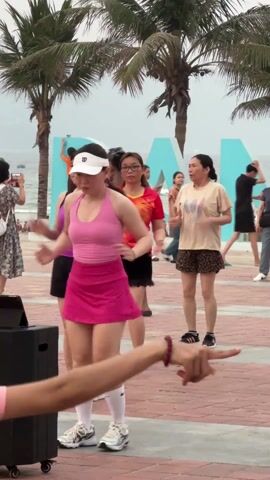 A group of women are participating in what appears to be an exercise class in a public outdoor setting. They are dancing or following an exercise routine. The women are of various ages and are wearing different colored outfits, including tank tops, shorts, and skirts. Some are wearing hats or visors. In the background, there are palm trees and a large sign that reads "DANANG." There are other people visible in the background, including a few men and women walking or standing around. The area where they are exercising appears to be a paved public plaza near the ocean. A hand with a red bracelet is briefly visible at the bottom of the screen, pointing towards the dancers. A black speaker is on the left side of the screen.