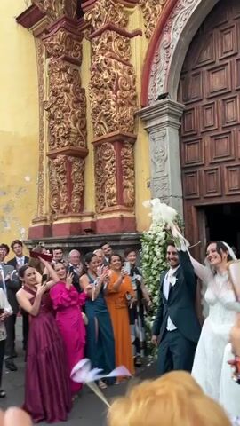 A wedding couple exits a church, arms raised in celebration, with guests cheering and throwing confetti. The bride wears a long-sleeved white lace gown and a long veil. The groom wears a dark green suit. The church has ornate architectural details with a yellow exterior. The crowd of guests includes people of varying ages, dressed in formal attire. Across the street, a building is visible with the words "HOSPITAL DE SALUD MENTAL ORIZABA" inscribed on its façade. Photographers and guests take pictures of the couple. The overall scene is celebratory and joyful.