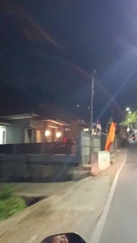 This media file appears to be a short video clip captured at night in Panembangan, Indonesia. The primary subjects are flags and a building with illuminated windows. An orange flag is prominently displayed, flapping in what seems to be a gentle breeze, positioned alongside a red and white flag, which could be the Indonesian flag.

The scene is set outdoors, along what looks like a paved area, possibly a sidewalk or a street. The time of day is clearly night, indicated by the dark sky and the artificial lighting emanating from the building and a street lamp visible in the background. The building itself has a somewhat modern, possibly residential or commercial, facade with multiple windows that are lit from within, suggesting occupancy. The architecture features textured walls and what appears to be a railing or fence in front.

There are no people visibly present in the clip. The overall mood is quiet and still, with the movement limited to the flags. The presence of flags, especially the national flag, might hint at a local celebration or a day of national significance. The setting appears to be a somewhat ordinary street scene at night. No discernible text is visible within the frame.