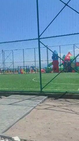 The video shows a view through a green chain-link fence at an outdoor recreational area on a sunny day. Immediately inside the fence is a green artificial turf soccer field with white markings. Beyond the soccer field is a playground area with brightly colored slides, climbing structures, and other play equipment. In the background, there are trees and a body of water that appears to be the ocean. The sky is clear and blue. There are no people visible on the soccer field or in the immediate playground area, although figures can be seen in the distance within the playground.