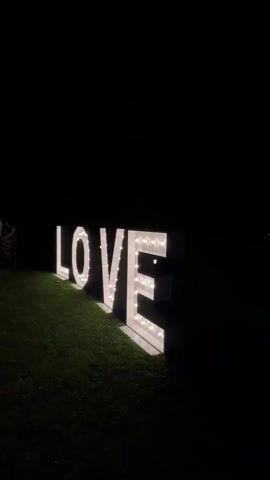 The video opens with a shot of a large, illuminated "LOVE" sign on a grassy area at night. The camera then pans to the left, revealing a covered outdoor event space with tables set for a gathering. The space is lit with various colored lights, predominantly green and blue, creating a festive atmosphere. Several people are visible inside the event space, some standing and some seated at the tables. The camera continues to pan across the interior, showing more tables and chairs, as well as what appears to be a DJ booth or entertainment area. The video ends by panning back towards the entrance of the event space.