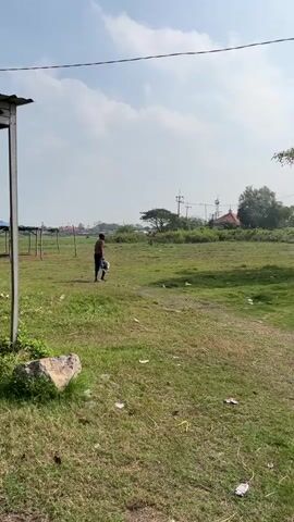 A person walks across a grassy field carrying an object. The field is littered with trash. In the background, there are trees, buildings, and utility poles. The sky is partly cloudy.