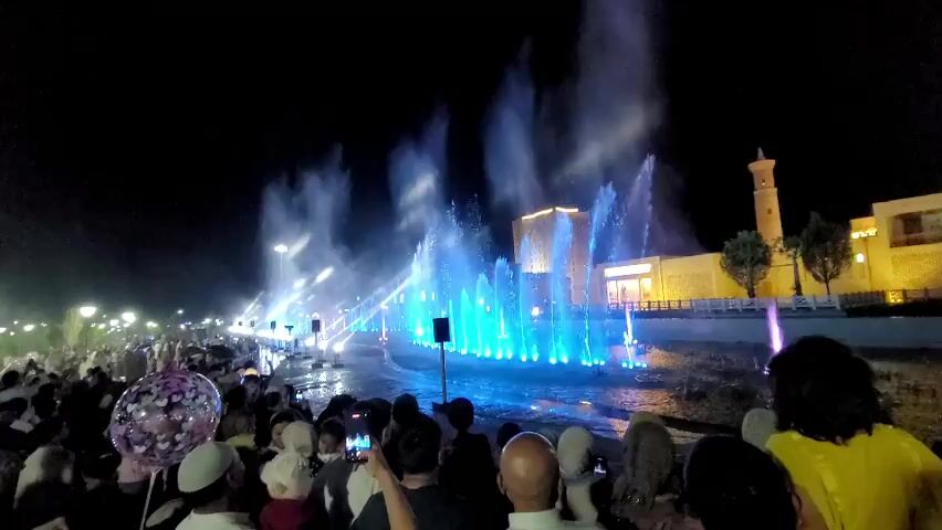 A nighttime aerial shot shows a large crowd of people gathered to watch a water fountain show. The fountains are illuminated with colorful lights, creating a dazzling display. The crowd is diverse, with people of all ages and backgrounds. Many people are holding up their phones to take pictures and videos of the show. The fountains are a mix of tall, powerful jets and smaller, more delicate sprays. The water is illuminated with a variety of colors, including blue, purple, and white. In the background, there is a building with a traditional architectural style, and some trees are visible. The overall atmosphere is festive and celebratory.