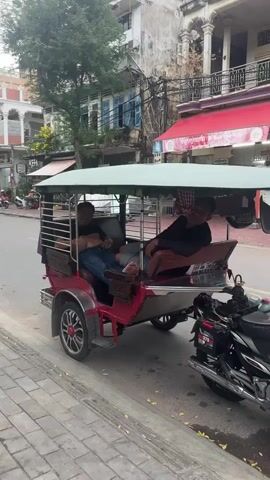 A tuk-tuk with two passengers is parked on the side of a street in front of buildings. One passenger, wearing a black t-shirt and blue jeans, is lying down in the back, his legs extended. The other passenger, also wearing a black t-shirt, is sitting on a bench in the front part of the tuk-tuk, leaning back and looking towards the camera. A third person, a woman with long brown hair and wearing a patterned dress, is walking by the tuk-tuk holding a smartphone. She is looking at the phone and then towards the people in the tuk-tuk. The people in the tuk-tuk appear to be interacting with the woman. A motorcycle is parked next to the tuk-tuk. The street is paved, and there are trees and buildings in the background.