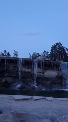 The video shows a wide, man-made waterfall cascading over a rough-hewn rock formation. The water is a clear, vibrant blue, and it falls into a dark, still pool at the base of the rocks. Above the waterfall, a dense forest of tall, green trees lines the horizon, their branches reaching towards a clear, pale blue sky. The lighting suggests it is twilight or early evening. The foreground is a rocky, sandy area, with large stones visible. The overall scene conveys a sense of natural beauty and tranquility.