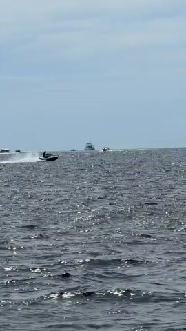 The video shows a personal watercraft moving across the water at high speed, leaving a wake behind it. A person is riding the watercraft. The water is dark and choppy, reflecting the sunlight. In the distance, a large ship is visible. The sky is pale blue with some clouds. The video ends with a close-up shot of a portion of a boat frame.