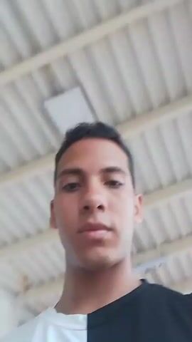 A young man wearing a black and white short-sleeved t-shirt is looking directly at the camera in a video that is shot from his perspective. The video begins with a close-up shot of his face, and then the camera slowly pans up towards the ceiling. The ceiling is white with several light fixtures, and a white sign with blue lettering can be seen in the background. The man's hair is dark and styled, and he has a serious expression on his face. He does not speak or move throughout the video. The video ends with the camera focused on the ceiling.