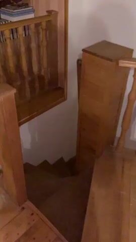 A wooden staircase descends from a landing. A wooden railing with vertical spindles lines the landing and a portion of the staircase. A tall wooden cabinet stands to the right of the staircase. The walls appear to be white.