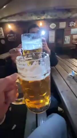 Two people are at an indoor bar, each holding a large mug of beer with a foamy head. One person is closer to the camera, holding their mug up. The other person, seated in the background, is taking a picture or video of the beer with their phone, which is emitting a bright light. The bar has wooden walls decorated with pictures and signs, and there are lights hanging above. The table is made of wood planks. The person in the foreground is wearing jeans. The overall lighting is dim, except for the light from the phone.