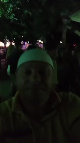 A first-person perspective selfie video shows a man wearing a yellow polo shirt and a white kufi. He is in a dimly lit, crowded outdoor setting, possibly a night market or festival, with other people visible in the background. The man smiles and makes a peace sign with his right hand, his arm raised. The background is blurry and illuminated by green and purple lights. The video is short, lasting only a few seconds.