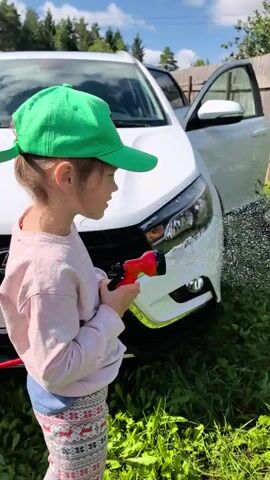 A young child wearing a green cap and a light-colored shirt is seen from the side holding a red and black garden hose with water spraying from it onto the front of a white car parked in a grassy area. The car has its driver's side door open. Behind the car is a weathered wooden fence and lush green grass and foliage. The video cuts between shots of the child with the hose and the water spraying against the fence.