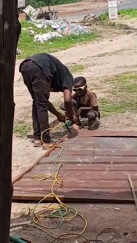 An adult male is using an angle grinder on a corrugated metal sheet. He is wearing a black shirt and dark pants. He is bent over the metal sheet, which is lying on the ground. A young boy is crouching nearby, wearing sunglasses and a patterned shirt. He appears to be watching the adult male. The scene is outdoors, with a dirt and gravel ground. There are some electrical cords on the ground, one yellow and one green. In the background, there is some vegetation and a pile of debris.