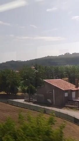 This video shows a view from a vehicle traveling along a road. In the foreground, there is a curved asphalt road with a grassy embankment on the left. Further back, a house with a red tile roof is visible, along with another building. Beyond the houses, there are trees and rolling hills. The sky is partly cloudy. The scene appears to be rural.