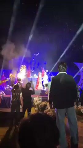 The video shows a wedding celebration at night. A bride and groom are standing on a stage decorated with flowers, illuminated by spotlights and fireworks, with people standing in front of the stage watching and taking photos. The groom is wearing a white suit, and the bride is in a red lehenga. The fireworks are large and colorful, creating a dramatic backdrop. The crowd is mostly blurred, but it appears there are several people in suits and formal wear.