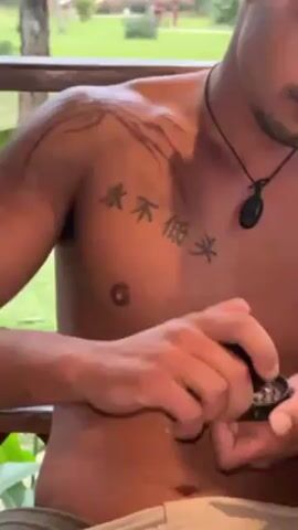 A shirtless man with tattoos on his shoulder and chest is handling a black container. He is on a porch overlooking a grassy area with trees and buildings in the background.