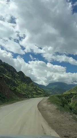 The video shows a scenic view from inside a vehicle driving on a winding road through a mountainous landscape. The road is paved and appears to be in fair condition, with white lane markings visible. On the left side of the road, steep, green, and rocky mountains rise sharply, covered in lush vegetation. The right side of the road transitions into a dirt shoulder and then slopes down into a valley. In the distance, more mountains are visible, some shrouded in clouds. The sky is a vibrant blue, with large, fluffy white clouds creating dramatic shadows and highlights across the landscape. The overall impression is one of natural beauty and vastness. The weather appears to be partly cloudy, with sunshine breaking through. The time of day seems to be daytime, likely late morning or early afternoon, given the bright lighting. The location is identified as Verkhnyaya Balkariya, Russia, which is known for its stunning Caucasian mountain scenery. There is no discernible activity or event taking place other than the act of driving through this beautiful environment. The mood evoked is one of peacefulness, adventure, and awe at the grandeur of nature. There is no visible text in the video.