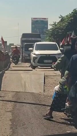 A group of people are on motorcycles on a bridge. In the foreground, a person is sitting on the back of a motorcycle, facing away from the camera. The motorcycle is parked. Behind this motorcycle, another motorcycle with two people on it is moving forward. A white car is approaching from the opposite direction on the bridge. Further down the bridge, other motorcycles and trucks are visible, suggesting traffic. Flags are mounted on the sides of the bridge, and a large banner is displayed in the background. The sky is clear and bright.