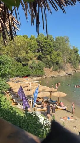 A sunny day at a crowded beach. The camera is looking down from above, with straw-like foliage in the foreground. Below, a sandy beach is lined with many sun loungers and thatched umbrellas. Several people are relaxing on the loungers, while others are walking on the beach. The sea is clear and blue, with numerous people swimming and wading in the water. Boats are anchored in the distance. Lush green trees and a rocky hillside form the backdrop to the beach. The sky is a clear, bright blue.