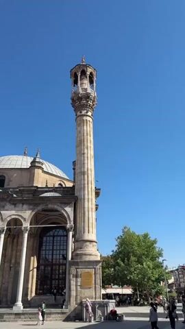 THE VIDEO SHOWS A LOW-ANGLE EXTERIOR SHOT OF A MOSQUE WITH A TALL MINARET. THE MOSQUE HAS A DOME AND A COLONNADED ENTRANCE. SEVERAL PEOPLE ARE VISIBLE IN THE FOREGROUND AND MIDGROUND, SOME WALKING, OTHERS STANDING. THE SKY IS CLEAR AND BLUE. TREES ARE VISIBLE IN THE BACKGROUND. THE VIDEO IS SLIGHTLY SHAKY.