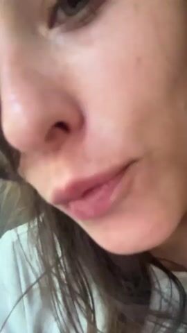 This media file features a close-up, selfie-style video of a young woman. She has long, dark, wavy hair that partially obscures her face, and light green eyes. She is wearing a white t-shirt with a red graphic design that appears to say "Book" or something similar in a stylized font. A silver necklace is visible. The lighting suggests it is daytime, with some harsh glare, possibly from sunlight.

The woman is looking directly at the camera and appears to be smiling or smirking. Her lips are slightly parted, and she seems to be in a relaxed or playful mood. The background is slightly out of focus, but some elements suggest an indoor setting, possibly a car or a room, with glimpses of what might be a window or another person in the far background.

The overall impression is of a casual, candid moment captured by the woman herself. There are no clear indications of a specific event, but the intimacy of the shot and her expression suggest a personal recording. The location context of Sokcho-si, South Korea, does not provide specific visual cues within the frame.