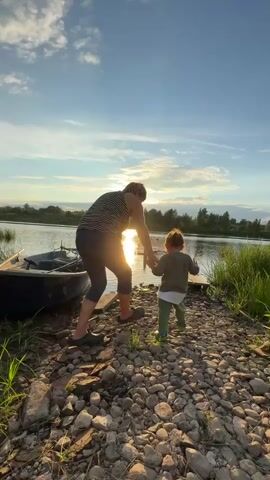 The video captures a rear view of a woman and a young child walking towards a small wooden dock on a rocky shore next to a calm body of water. The sun is low in the sky, casting a warm glow and creating a silhouette of the woman. A small blue boat is moored to the left of the dock. Lush green grass lines the edge of the water. The sky is clear with a few wispy clouds. The woman is wearing a striped top and dark pants, while the child is dressed in a green top and light-colored pants. The woman's hand is on the child's back, guiding them towards the dock. The scene suggests a peaceful moment by the water during sunset.