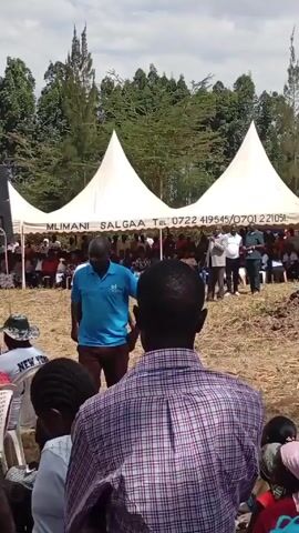 The video depicts an outdoor gathering. A large group of people are seated under white tents. A man is standing in front of the crowd and appears to be speaking. The crowd is mostly looking at the speaker. Several trees are visible in the background. The ground is mostly dirt. The camera is positioned behind several people in the audience.