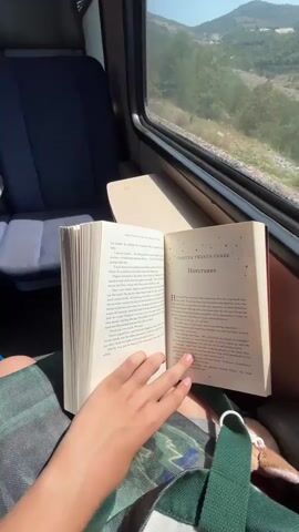 A close-up shot shows a person's hands holding open a book titled "Harry Potter and the Half-Blood Prince" while sitting on a train. The book is open to chapter twenty-three, "Horcruxes." Through the train window, a scenic view of rolling hills and a body of water is visible under a clear blue sky. The train tracks run alongside the window, and power lines are visible in the distance. The lighting suggests it is daytime, likely during sunny weather.