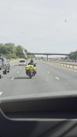 The video shows a view from inside a vehicle traveling on a multi-lane highway. In the foreground, the top portion of the vehicle's dashboard is visible. The highway is paved and marked with white lane lines. In the middle distance, a motorcycle with a yellow bag attached to the rear is traveling in the right lane. There are other vehicles on the highway, including cars and what appears to be a white van. An overpass spans the highway in the background, and trees and vegetation line the sides of the road. The sky is visible above, with some clouds.