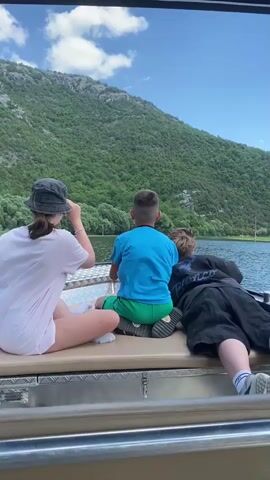 The video shows three people from behind, seated on a boat. The person on the left is wearing a light pink t-shirt and a dark bucket hat. The middle person is a child wearing a bright blue t-shirt and green shorts, sitting on a dark cushion. The person on the right is wearing a dark t-shirt and black shorts. They are all looking out at a body of water, with a lush, green, tree-covered mountain rising in the background under a bright blue sky with scattered clouds. The water appears calm. The scene suggests a leisurely boat ride on a sunny day, likely in a scenic, natural setting like the one described in Meterizi, Montenegro. The overall mood seems peaceful and observational, as the subjects are taking in the view. There is no visible text in the video.
