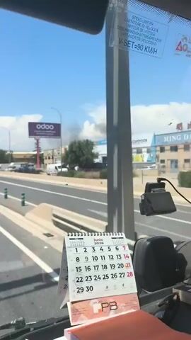 The video shows a view from inside a vehicle looking out at a street scene. A large plume of black smoke is visible rising into the sky in the distance, indicating a fire. Several buildings and signs, including one for "odoo," are visible along the street. Traffic is moving on the road, with cars and vans. A calendar showing June 2020 is visible on the dashboard of the vehicle. The sky is blue with some clouds.
