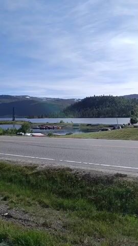 This media file captures a scenic view of a lake and surrounding landscape on a sunny day, likely in Hovden, Norway. The scene is dominated by a calm body of water reflecting the bright blue sky, which is adorned with wispy white clouds. In the background, rolling hills covered in dense green forests rise up, their peaks gently touched by snow, suggesting a high-altitude or northern location.

On the left side of the lake, a tall, dark structure, possibly a tower or an antenna, stands prominently against the horizon. Along the shoreline, several small boats and kayaks are visible, some pulled onto the grassy bank, hinting at recreational activities like boating or fishing. A paved road runs horizontally across the foreground, with a strip of lush green grass and vegetation bordering it. Further back, near the lake, a parking area with several cars is present, indicating that this is a location accessible to visitors. A few trees are scattered throughout the landscape, adding to the natural beauty.

While no specific individuals are clearly identifiable as the main subjects, there appear to be a few people in the distance near the parked cars and by the lake, suggesting a peaceful, leisurely day. The overall mood conveyed is tranquil and serene, bathed in the bright, clear light of what appears to be midday or early afternoon. The presence of both natural beauty and amenities like a road and parking area suggests this is a popular spot for outdoor recreation and enjoying the Norwegian wilderness. No visible text is present in the media file.
