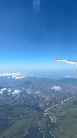 The video presents an aerial view from an airplane window, showcasing a vast mountainous landscape under a clear blue sky. On the right side of the frame, a portion of the airplane's white wing with a prominent winglet is visible, indicating the flight's altitude.

Below the aircraft, an extensive range of rugged, greyish-green mountains stretches across the horizon. These mountains are characterized by their deeply carved valleys and undulating terrain. A winding river, appearing light brown or sandy, snakes its way through one of the prominent valleys, cutting through the elevated land. Scattered white clouds dot the sky, some floating below the airplane, and others further away, adding depth to the scene. The overall weather appears clear and sunny, providing excellent visibility of the terrain.

The scene captures the serene and expansive beauty of the landscape as seen from above, suggesting a journey over remote and majestic natural formations in Arzikush, Azerbaijan. No people or visible text are present in the frame.