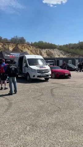 Several people walk around a parking lot on a sunny day. Parked in the lot are a variety of sports cars, including McLarens, a Porsche, and a Toyota Supra. A white van is also parked nearby. In the background, there is a hill and a couple of portable structures.