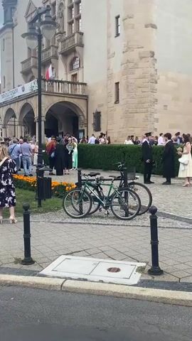 The video begins with a slightly low-angle, shaky shot of a building's exterior and a paved courtyard. Two bicycles are parked prominently in the foreground, facing left. The building appears to be made of light-colored stone, with an arched entryway, a balcony with a railing, and windows. A white flag with a red stripe is visible on a pole on the balcony. 

A group of people are gathered in the courtyard, some standing in a line near the building's entrance, while others are mingling. Many are wearing formal attire, including some in graduation caps and gowns. A few people are holding bouquets of flowers. A hedge runs along the front of the building, separating the gathered people from the paved area. 

The camera pans slowly to the right, showing more of the building and the crowd. In the middle ground, two individuals in graduation gowns are facing each other, appearing to interact. One is holding a bouquet. A person in a uniform, possibly a security guard or official, stands nearby. 

The camera continues to pan right, revealing more people dressed formally. A woman in a yellow dress and another in a white dress are visible. Further right, a couple stands facing away from the camera, the woman wearing a colorful sari. 

The video continues to show more of the crowd and the building's architecture. A man in a white shirt and jeans is visible on the far right. The camera briefly focuses on a drain cover in the foreground before the video ends. The overall impression is of a formal gathering or event taking place outside a historical building.