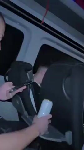 The interior of a passenger van is shown in a short video clip. Several individuals are seated, though only their backs and profiles are fully visible. One young man with short, dark hair looks directly at the camera from behind the seat in front of him and smiles. The camera holder is seen from the shoulders down, manipulating a white water bottle in their hands while holding a smartphone. Blue ambient lighting is present on the ceiling of the van. Tray tables are visible on the back of the seats. The environment appears to be dark outside, suggesting nighttime.