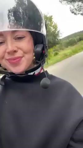A woman wearing a shiny, full-face helmet and a black sweater is recording a selfie video while riding some sort of vehicle, likely a moped, down a paved road. She appears to be smiling and sticking her tongue out briefly at the camera. The surrounding environment consists of green trees and vegetation. The sky appears overcast.