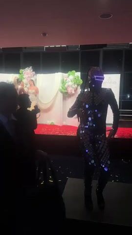 The video shows a backstage area, possibly for a beauty pageant or formal event. Three women in formal dresses, one with a sash, are posing for a photograph on a small stage with a red carpet. The backdrop includes white panels decorated with floral arrangements. A photographer is taking their picture, and there are silhouettes of other people in the foreground, including a figure covered in reflective discs who is dancing. Confetti is scattered on the floor.